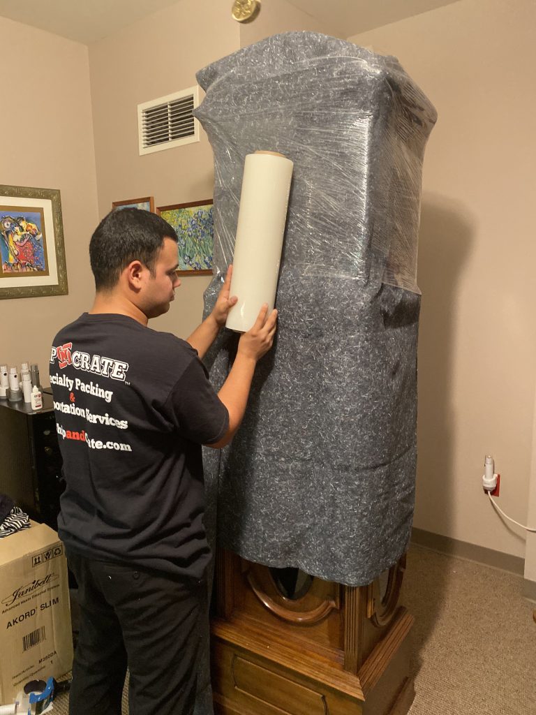 Ship-n-Crate packer shrink-wrapping a blanket-padded antique grandfather clock for long-distance shipping
